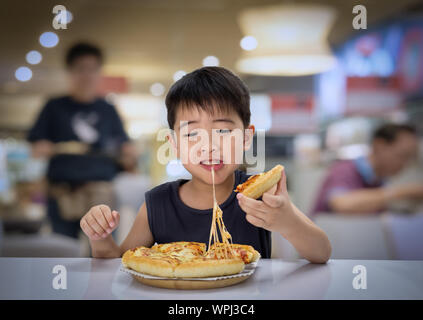 Asian 6-7 year boy is happy to eating pizza with a hot cheese melt stretched on a wooden pad in resturant. Stock Photo