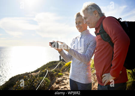 Smiling senior couple taking selfie while standing by sea against sky during sunny day Stock Photo