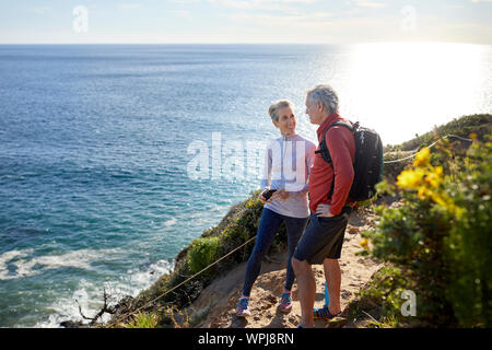 Full length smiling senior couple talking while standing on cliff by sea during sunny day Stock Photo