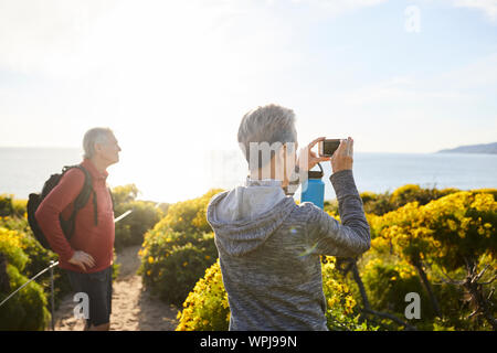 Rear view of senior woman photographing sea through smart phone while standing by plants against sky Stock Photo