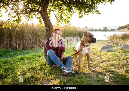 redhair jouful young woman caressing their dog, wearing sport clothing, enjoying their time and vacation in sunny park Stock Photo