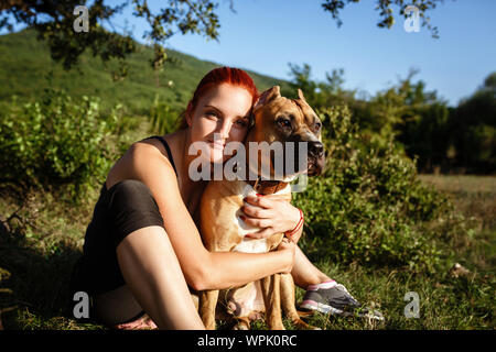redhair jouful young woman caressing their dog, wearing sport clothing, enjoying their time and vacation in sunny park Stock Photo