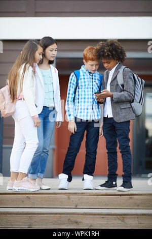 Group of friends with backpacks using mobile phone they standing outdoors near the school building and waiting for the lessons Stock Photo