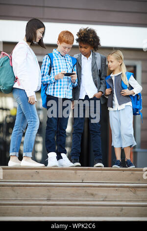 Group of students standing and looking at their classmate how he playing the game on mobile phone near the school building outdoors Stock Photo