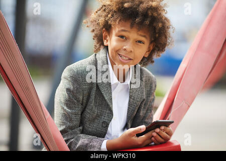 Portrait of African schoolboy using his mobile phone and smiling at camera while sitting outdoors in the park Stock Photo