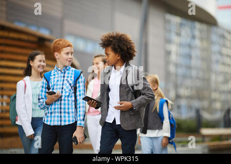 Two boys with backpacks holding mobile phones and talking to each other while walking along the street with other children in the background Stock Photo