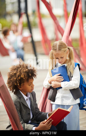 Two school classmates reading book and working together in team while sitting outdoors at schoolyard Stock Photo