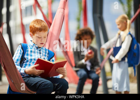 Red haired schoolboy resting after lessons and reading a book at schoolyard with other school children in the background Stock Photo