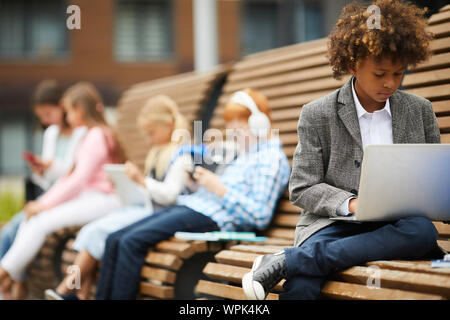 African schoolboy sitting on the bench and using laptop computer for study with his classmates sitting in the background Stock Photo