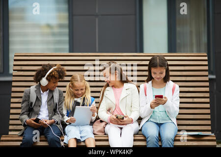 Group of school children sitting on the bench outdoors and using tablet pc and mobile phones they communicating online Stock Photo