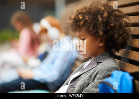 Side view of African boy with curly hair sitting on the bench outdoors and looking sad Stock Photo