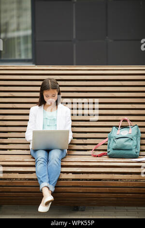Asian schoolgirl with backpack sitting on the bench and communicating online using her laptop computer outdoors Stock Photo