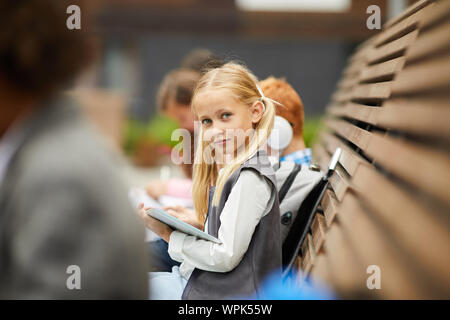 Portrait of schoolgirl with long blond hair looking at camera while sitting on the bench with her classmates and using digital tablet outdoors Stock Photo