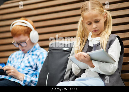 Cute schoolgirl with long blond hair using digital tablet for her study while sitting on the bench with schoolboy in the background Stock Photo