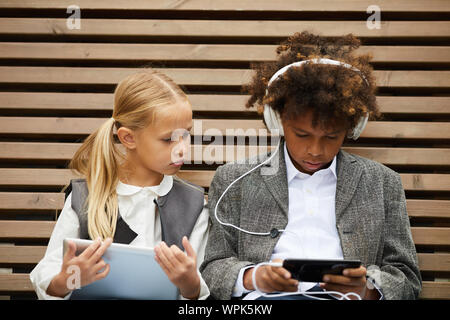 African schoolboy in headphones listening to music on his mobile phone with schoolgirl using tablet pc they sitting on the bench outdoors Stock Photo