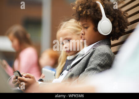 African boy with curly hair wearing headphones and playing online game on his mobile phone with girl sitting near by him on the bench outdoors Stock Photo