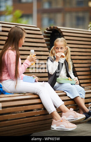 Two friends eating sandwiches and drinking juice while sitting on the bench together outdoors during school break Stock Photo