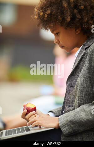 African serious boy with curly hair sitting and typing on laptop computer and eating fruit outdoors Stock Photo