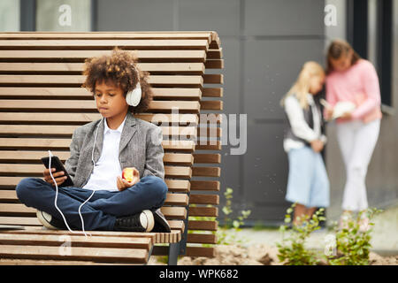 African boy with curly hair in headphones resting on the bench listening to music on mobile phone and eating fruit outdoors Stock Photo