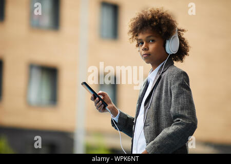 Portrait of African boy with curly hair wearing headphones and using his mobile phone while standing in the city Stock Photo