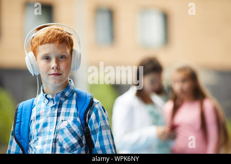 Portrait of red haired schoolboy with backpack looking at camera while wearing headphones and listening to music outdoors Stock Photo