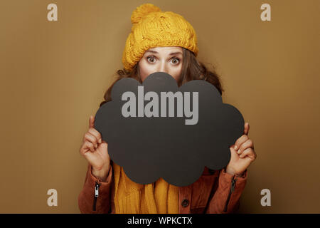 Hello autumn. surprised stylish woman in yellow beret and scarf hiding behind cloud shape blank blackboard against brown background. Stock Photo