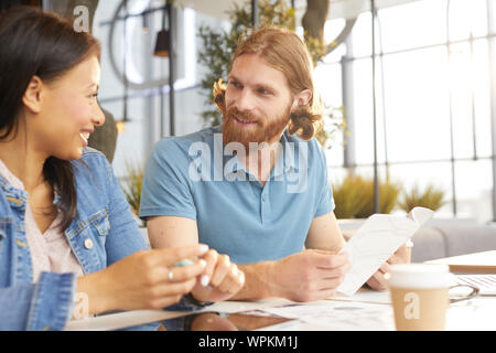 Red haired man sitting at the table with document and talking to young smiling woman while they have a meeting in cafe Stock Photo