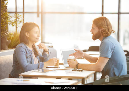 Two business colleagues in casual clothing sitting at the table with laptop drinking coffee and discussing new project together during meeting in cafe Stock Photo