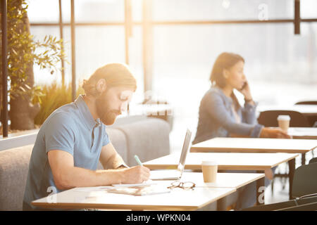 Serious bearded businessman sitting at the table with laptop and making notes with businesswoman sitting in the background in cafe Stock Photo