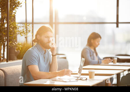 Serious bearded businessman in casual clothing talking on mobile phone while working at the table with laptop with woman in the background in cafe Stock Photo