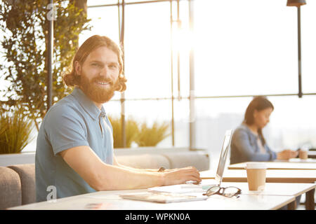 Portrait of successful bearded businessman smiling at camera while typing on laptop at the table in coffee shop Stock Photo