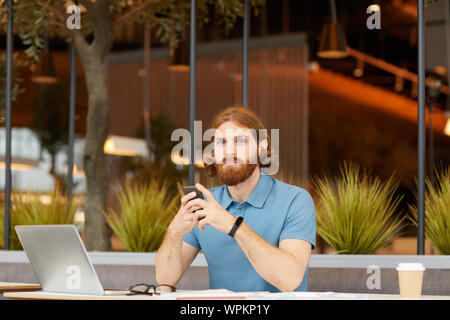 Portrait of red haired bearded man in casual clothing sitting at the table in front of laptop looking at camera and using mobile phone Stock Photo