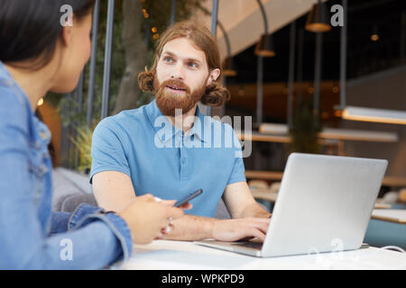 Young bearded businessman typing on laptop computer and talking to young woman while she using mobile phone during their meeting in cafe Stock Photo