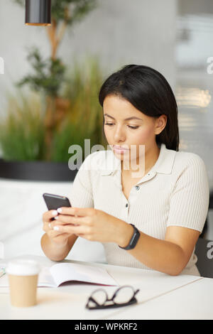 Young pretty brunette sitting at the table typing a message on mobile phone while drinking coffee in cafe Stock Photo