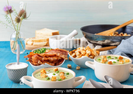 Blue wooden table with bowls of thick green pea soup embellished with roasted rusks, green peas, slices of grilled bacon, crushed black pepper and cre Stock Photo