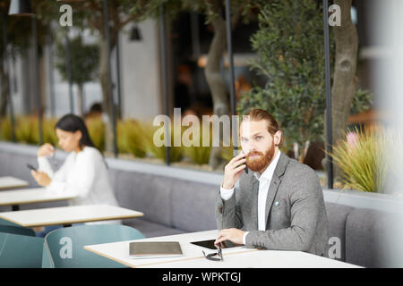 Portrait of bearded young businessman in formalwear sitting at the table and talking on mobile phone while waiting for his colleague at the cafe Stock Photo