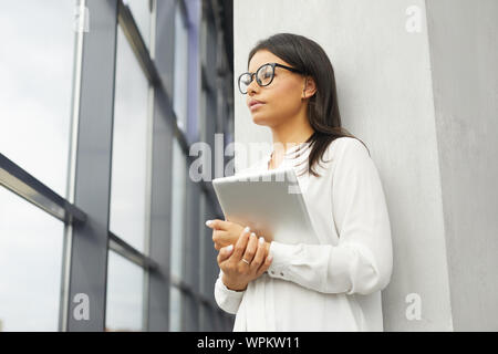 Young beautiful businesswoman in eyeglasses holding tablet pc and looking through the window with pensive sight while standing at office Stock Photo