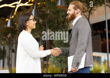 Two young business partners shaking hands to each other and smiling while standing outdoors in the city Stock Photo