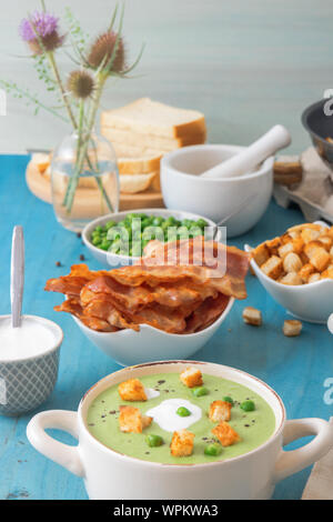Blue wooden table with bowls of thick green pea soup embellished with roasted rusks, green peas, slices of grilled bacon, crushed black pepper and cre Stock Photo