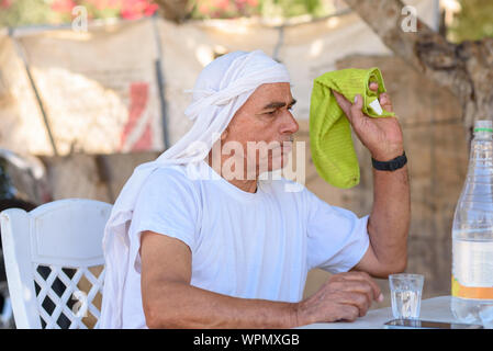 Bedouin senior tries to swat fly with towel. Old Arabic man in traditional clothes outdoor portrait. Elderly muslim men drinking water and sitting at a table chasing away the flies and insects. Stock Photo
