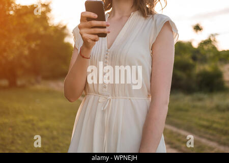 Beautiful young woman with brown hair in white dress walks through the autumn or summer forest with smartphone in her hand. Girl is looking for mobile Stock Photo