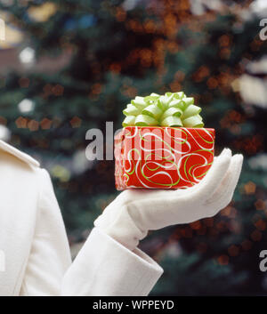 Woman's hand holding Christmas gift present wrapped in festive wrapping paper and bow. Holiday giving gifts presents background. Stock Photo
