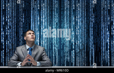 Man with folded hands looking upward. Young businessman sitting at desk. Programmer wears business suit and tie on background cyberspace with digital Stock Photo