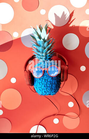 Funny pineapple painted blue in red in sunglasses and earphones. Flat lay on coral color paper with circles. Stock Photo