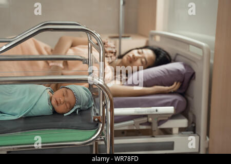 Newborn baby laying in crib with his mother lying on bed side Stock Photo