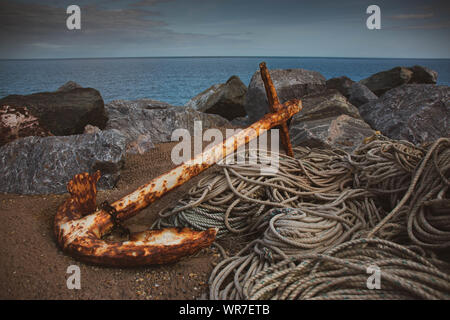 A rusty anchor and rope set on the rocks by the sea Stock Photo