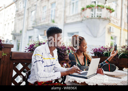 Handsome stylish african american couple sitting at outdoor cafe with laptop and making selfie on mobile phone. Stock Photo