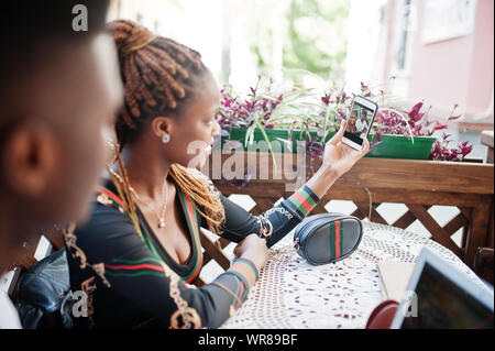 Handsome stylish african american couple sitting at outdoor cafe with laptop and making selfie on mobile phone. Stock Photo