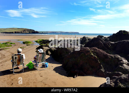 A couple enjoy the sun as they sit  on the beach at Bigbury-on-Sea, South Devon where there is access to Burgh Island Hotel and the Pilchard Inn. Stock Photo