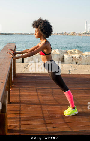 happy young african woman stretching and warming up before an outdoor workout. The Mediterranean sea in the background